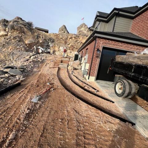 Rutted muddy road beside a brick house, with rocky hillside and a truck trailer on the right
