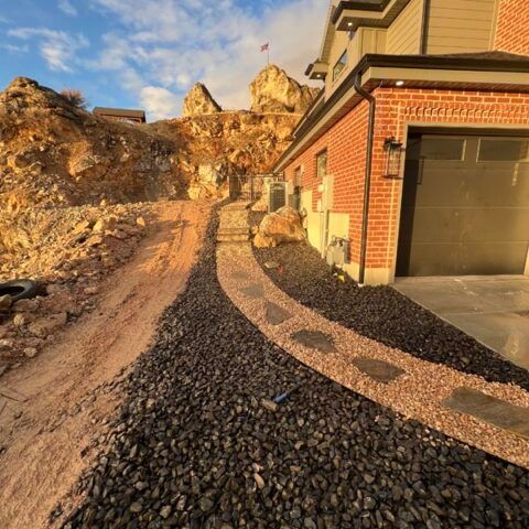 New brick house with garage beside a curved gravel driveway at sunset, backed by rocky hills