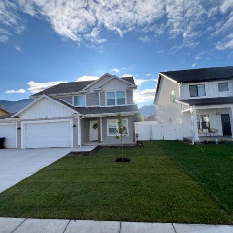 Suburban two-story house with attached garage, green lawn, and neighboring home under a bright blue sky