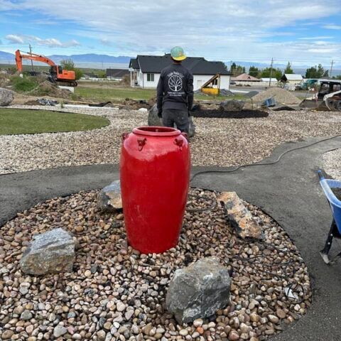Person standing near a bright red barrel in a landscaped construction area with rocks and excavator in background