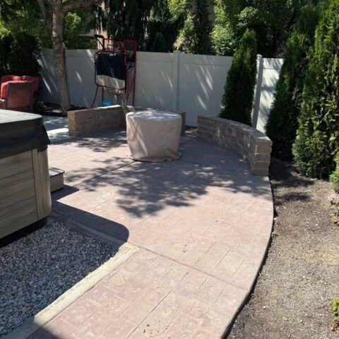 Curved stone patio with outdoor seating, a fire pit, and tall trees along a privacy fence.
