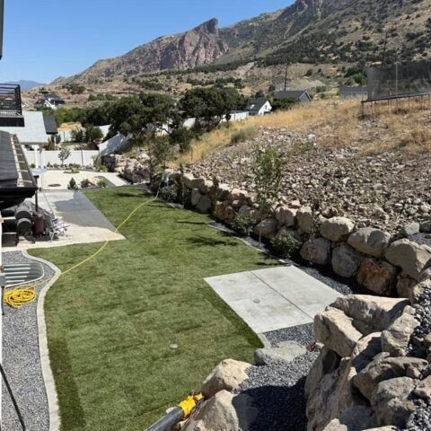 Rocky hillside yard with green lawn, stone retaining wall, and mountain backdrop under a clear blue sky