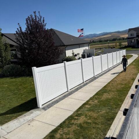 Suburban sidewalk beside a long white fence, with a person walking and mountains in the distance.