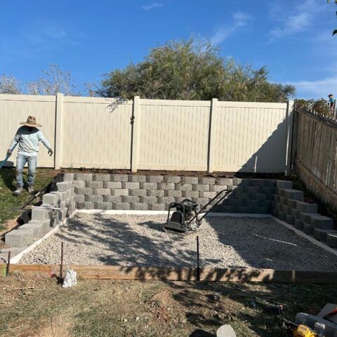 Backyard with retaining wall, gravel area, beige fence, and a worker on the left.