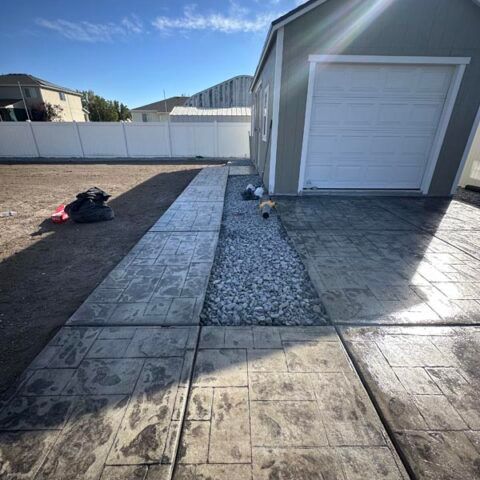 Backyard with new concrete walkway and gravel strip beside a gray garage and white fence