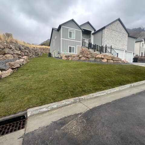 Grassy sloped yard beside a gray house and retaining rocks along a curb on a cloudy day