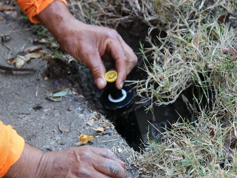 Hands adjust a black sprinkler valve in a narrow trench beside grass and concrete.