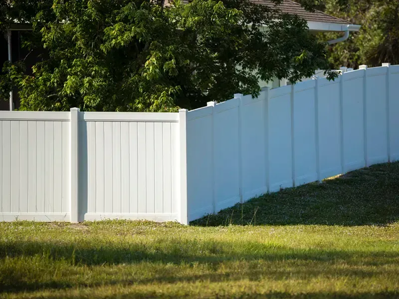 White vinyl fence along a grassy yard, with trees in the background and sunlight casting shadows.