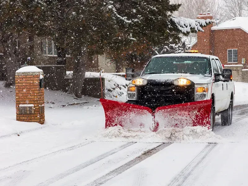 Pickup truck with red snowplow clearing a snowy residential street during snowfall