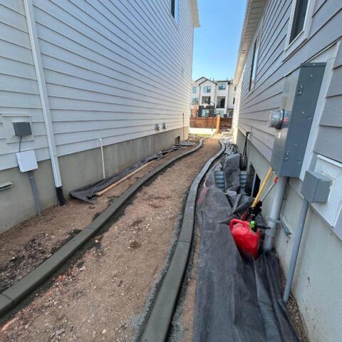 Narrow gravel walkway between two houses with black landscape fabric, boards, and red shovel near the side of a home