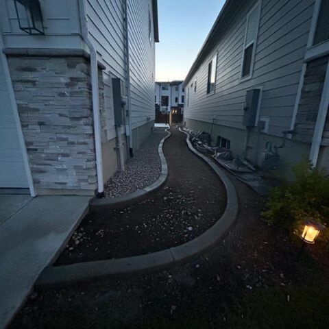 Narrow gravel path between two gray apartment buildings at dusk, with curved edging and a small garden light.