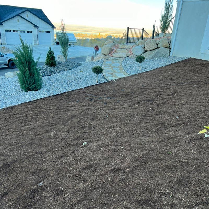 Front yard with new gravel landscaping, rocks, shrubs, and a dark mulch slope beside a house at sunset