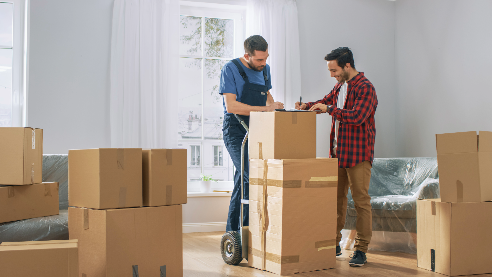 Two movers packing boxes in a bright room, sealing a large cardboard box on a hand truck.