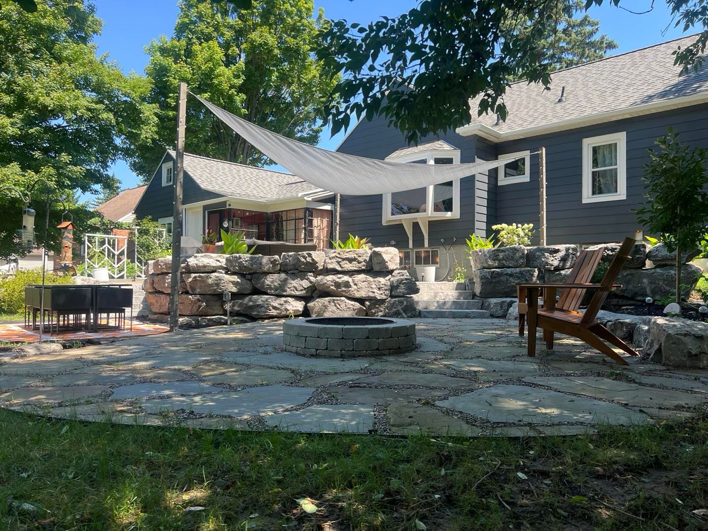 Backyard patio with flagstone, fire pit, and rock wall, under a white shade sail, beside a dark house.