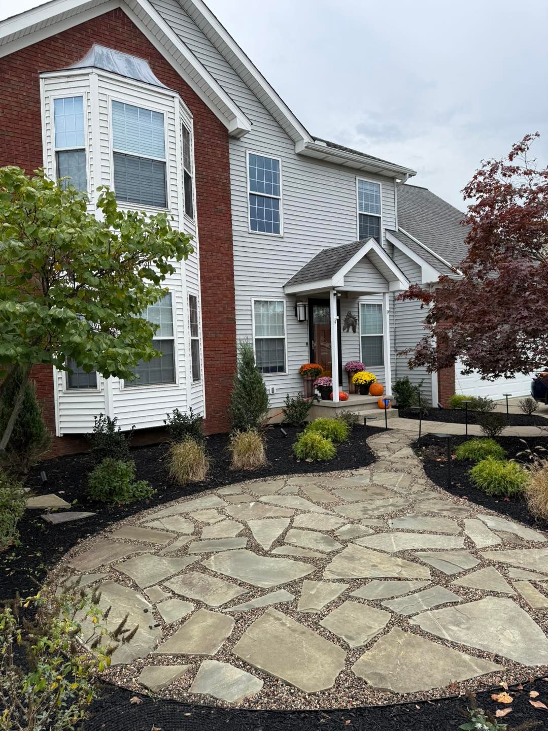 Stone walkway leads to a two-story house with brick and gray siding. Autumn decorations on porch.