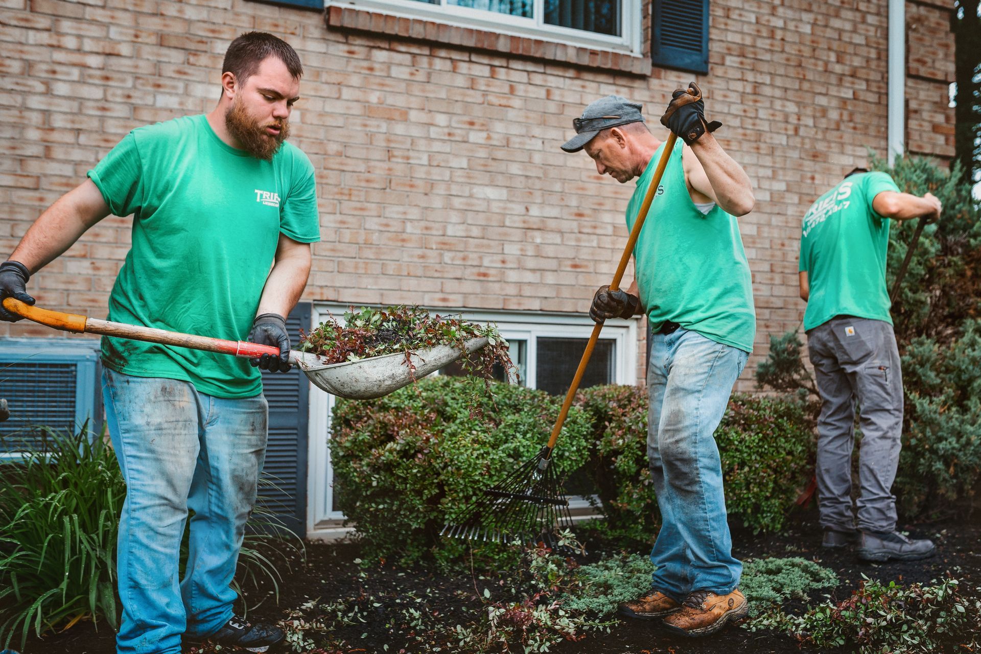 Three people in green shirts tending a garden bed in front of a brick building; one uses a shovel.