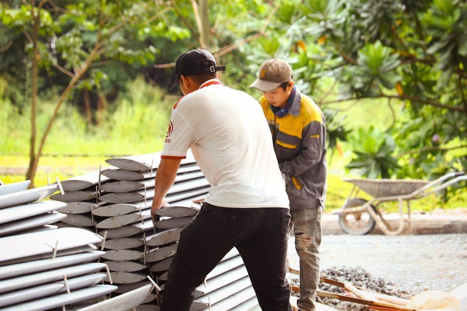 Two people lifting metal panels outside. One wears white shirt, the other a jacket. A wheelbarrow sits nearby.