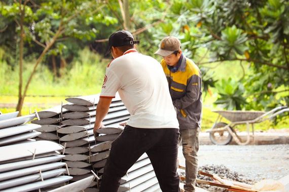 Two people lifting metal panels outside. One wears white shirt, the other a jacket. A wheelbarrow sits nearby.