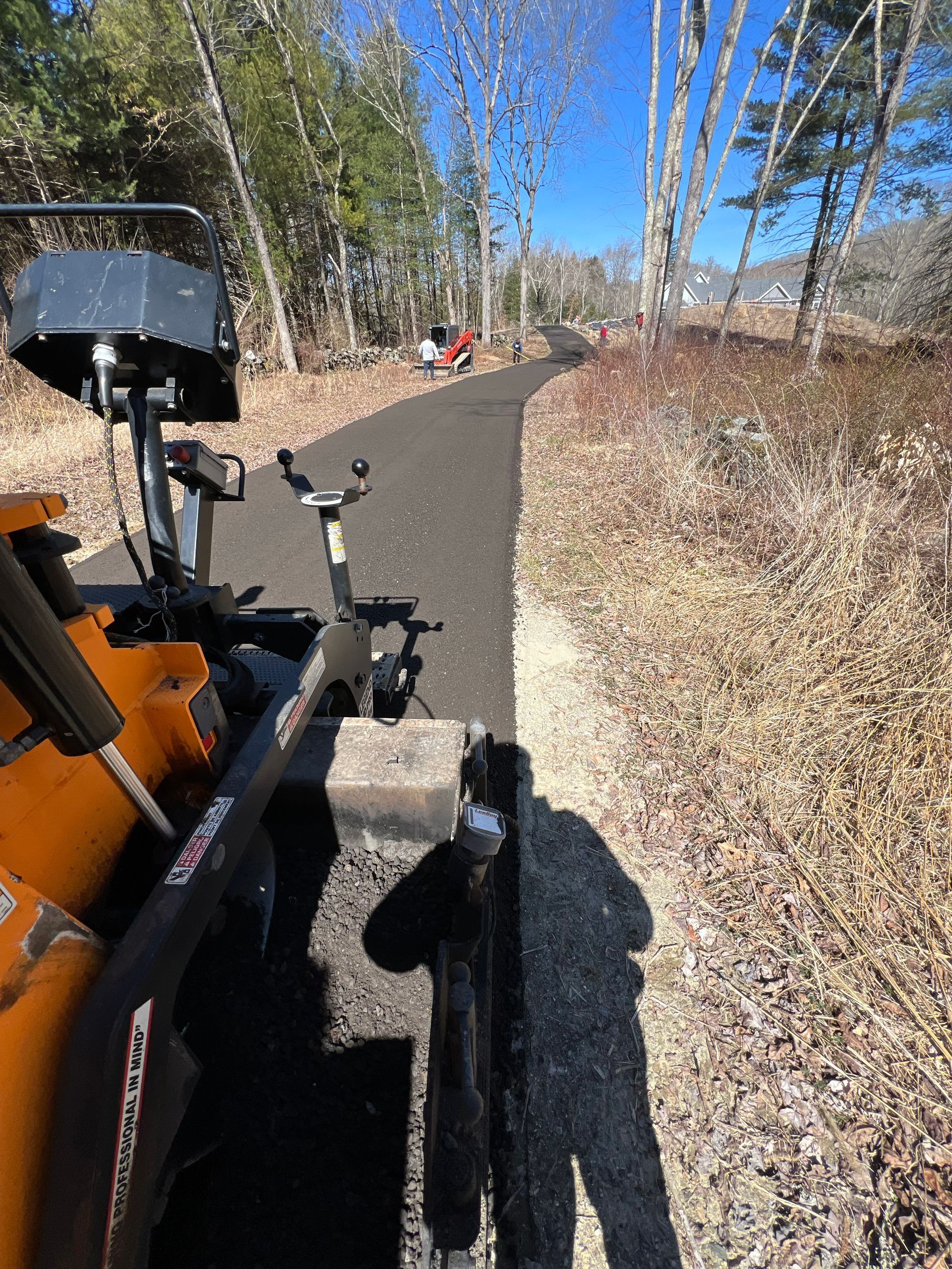 Paved walking path through woods with construction equipment visible on the left and workers in the distance.