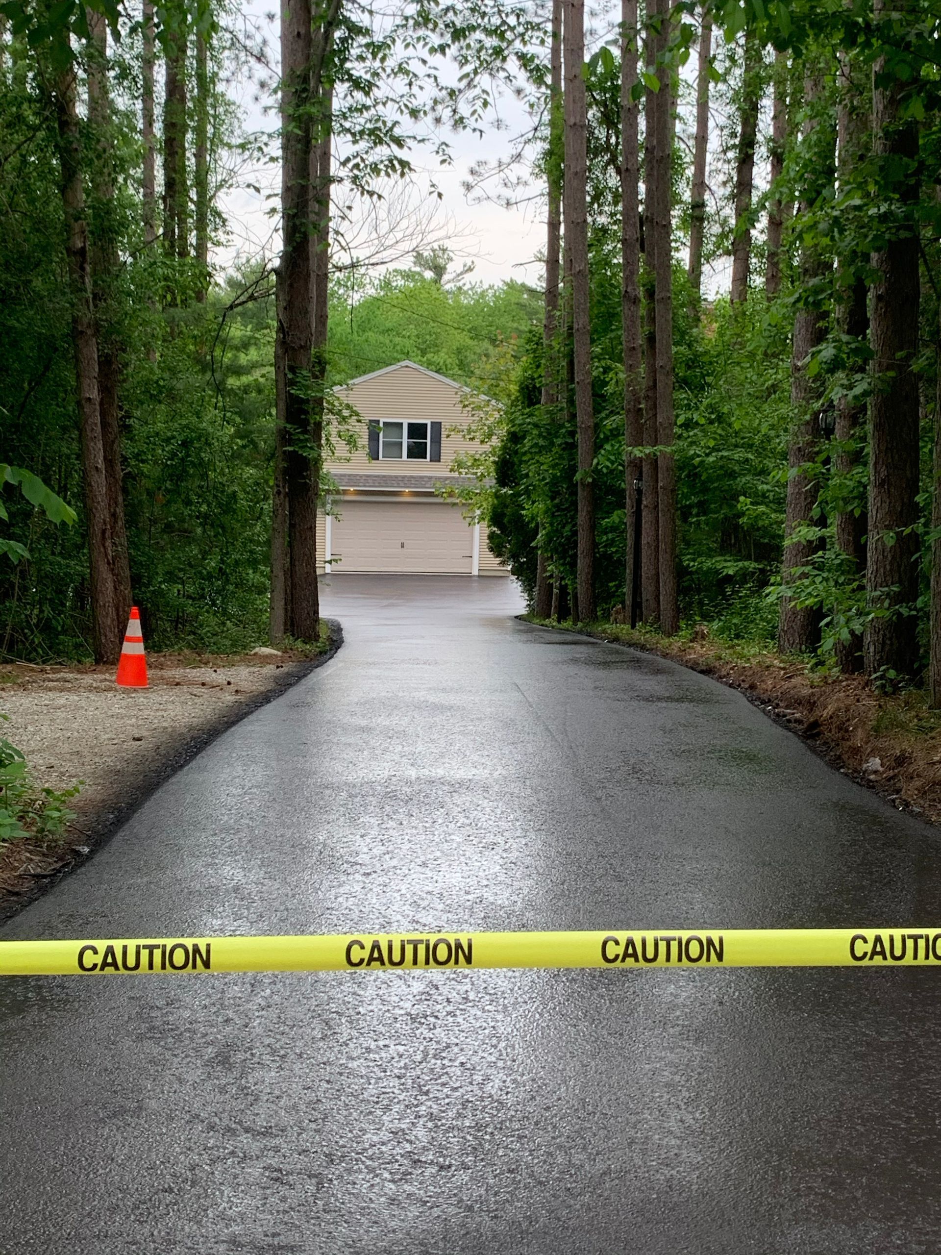 Black asphalt driveway leading to a house, blocked by yellow caution tape. Trees frame the scene.