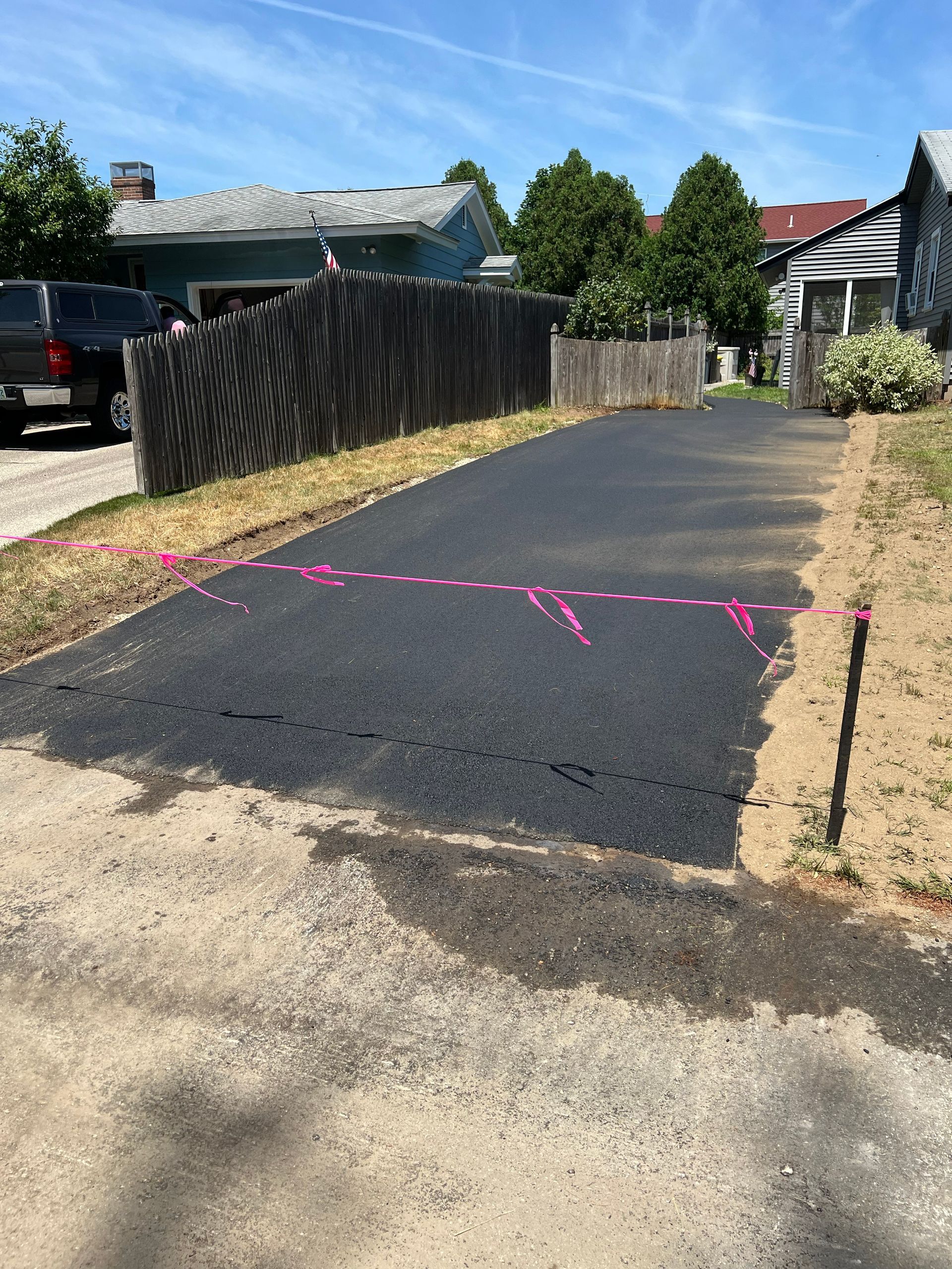 Freshly paved asphalt driveway with pink markings; residential setting with wooden fence and houses.