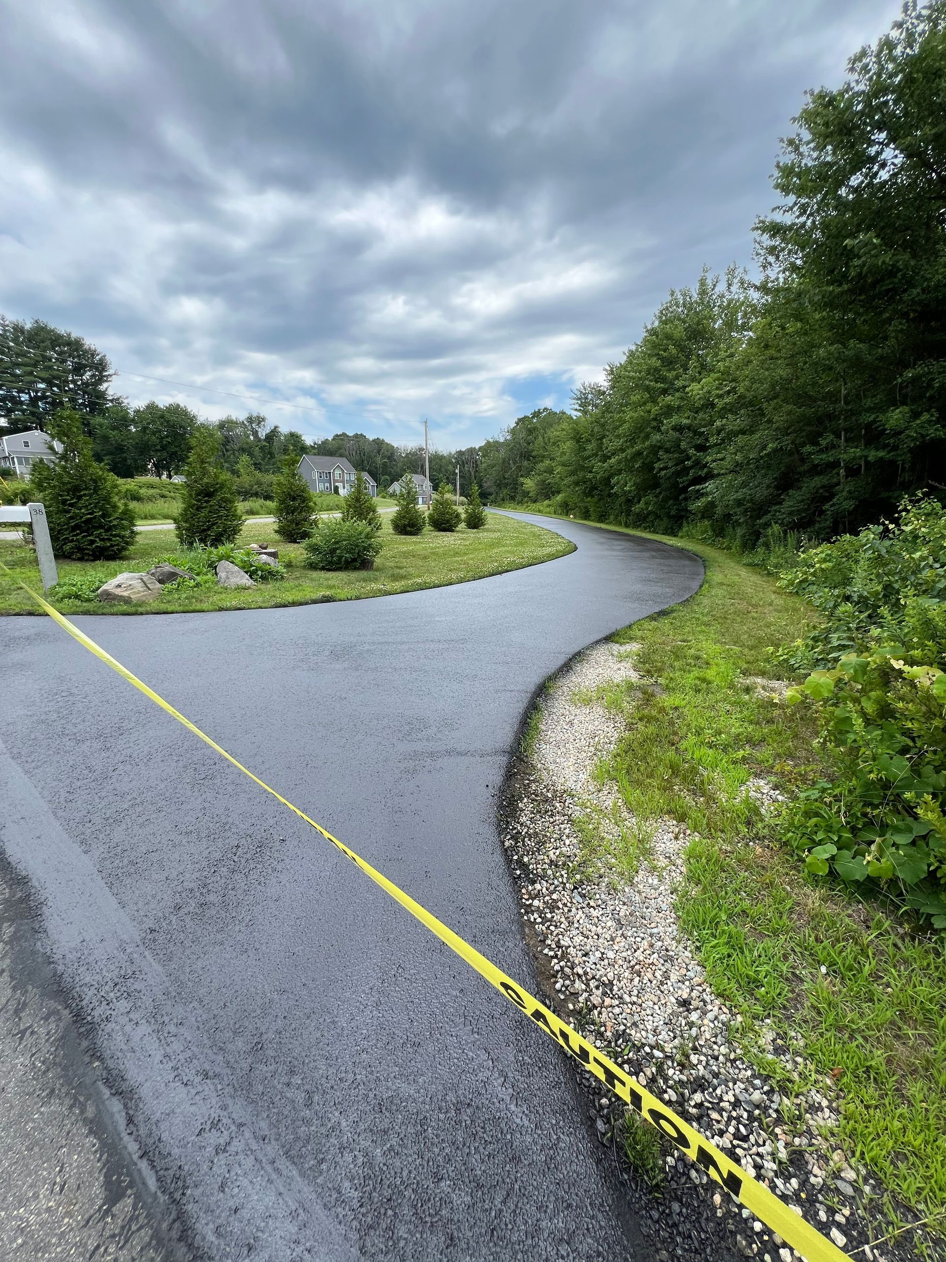 Newly paved road curves through a grassy area, with caution tape across the foreground. Cloudy sky.