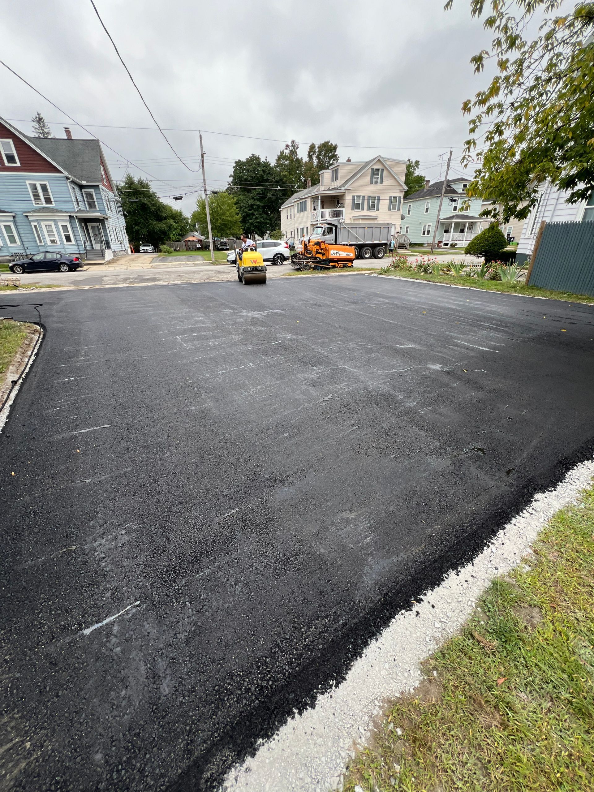 Newly paved asphalt street with a roller compacting the surface. Construction vehicles and houses are visible.