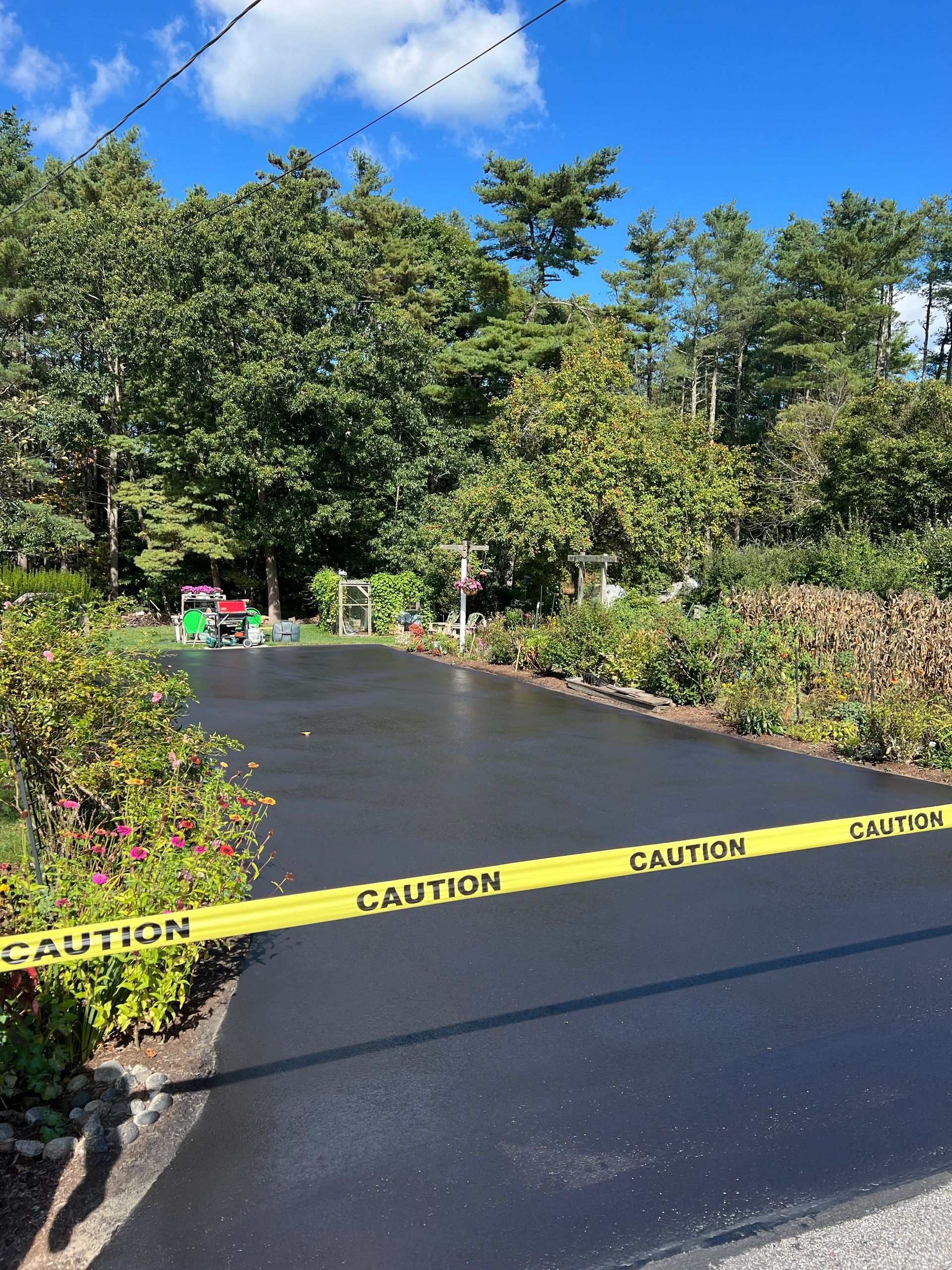 Newly paved driveway with yellow caution tape, lined by plants and trees. Bright, sunny day.
