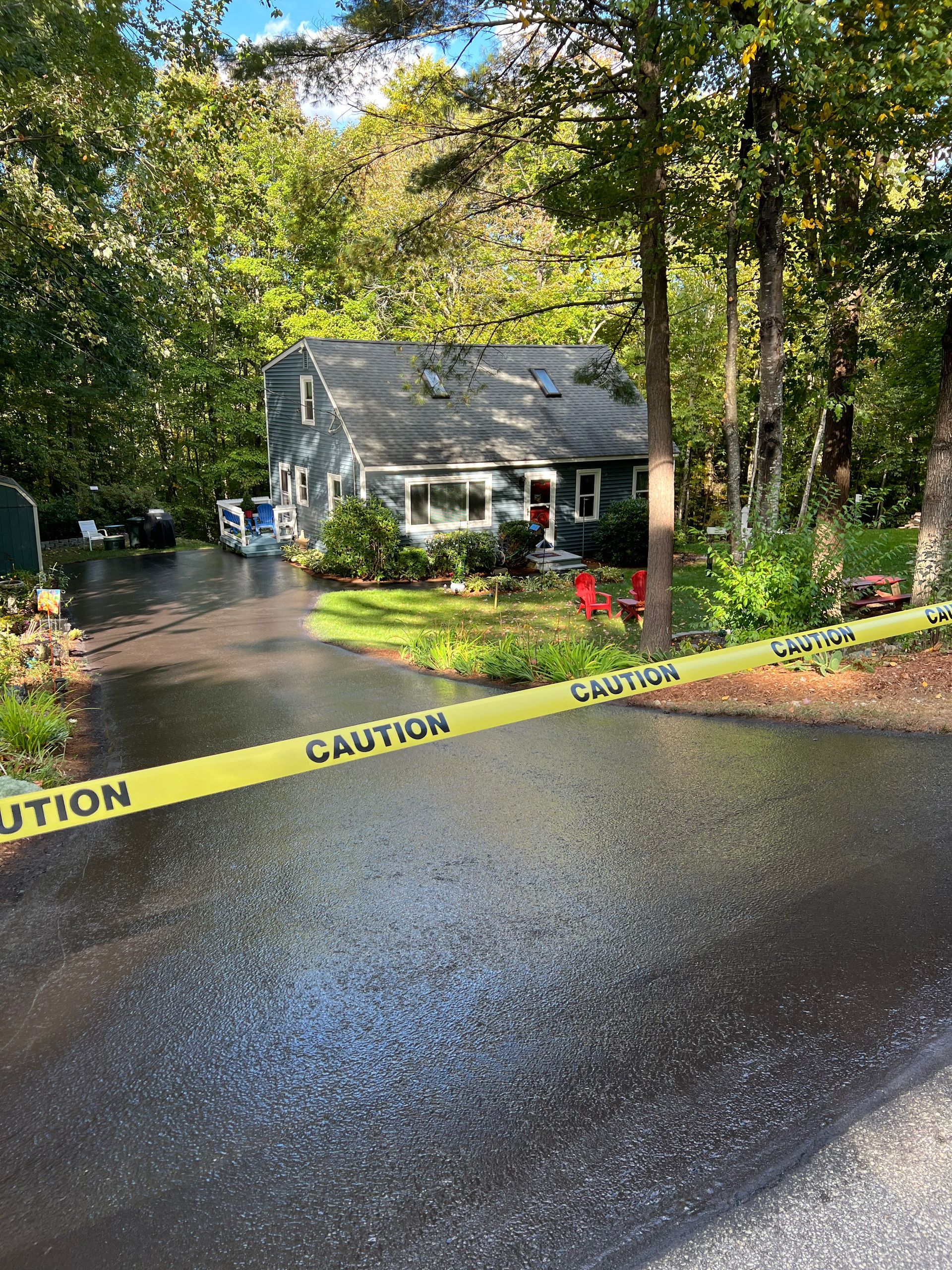 A gray house with caution tape in front. Wet asphalt driveway, surrounded by trees.