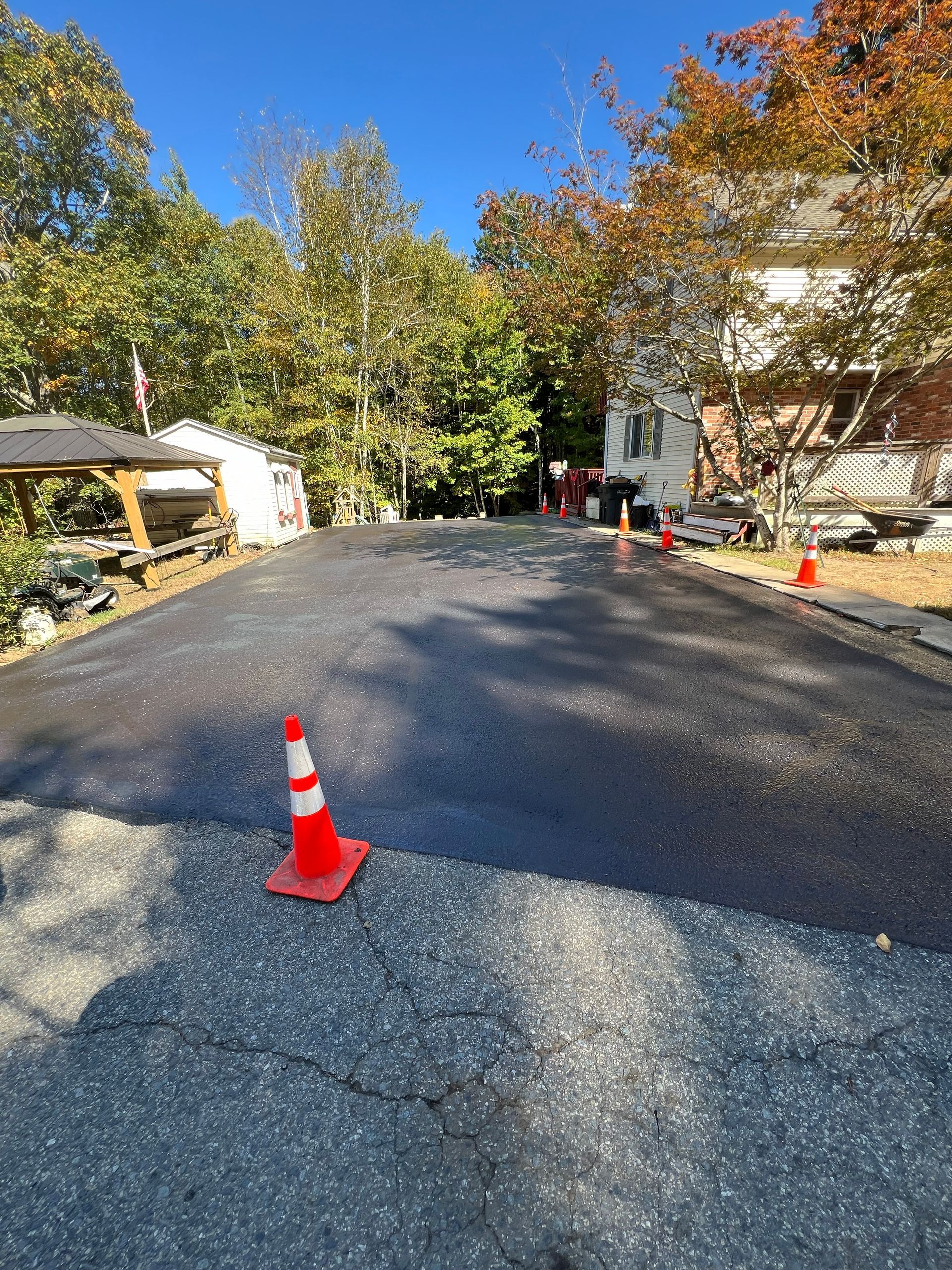 Newly paved asphalt driveway with orange traffic cones and trees in the background.