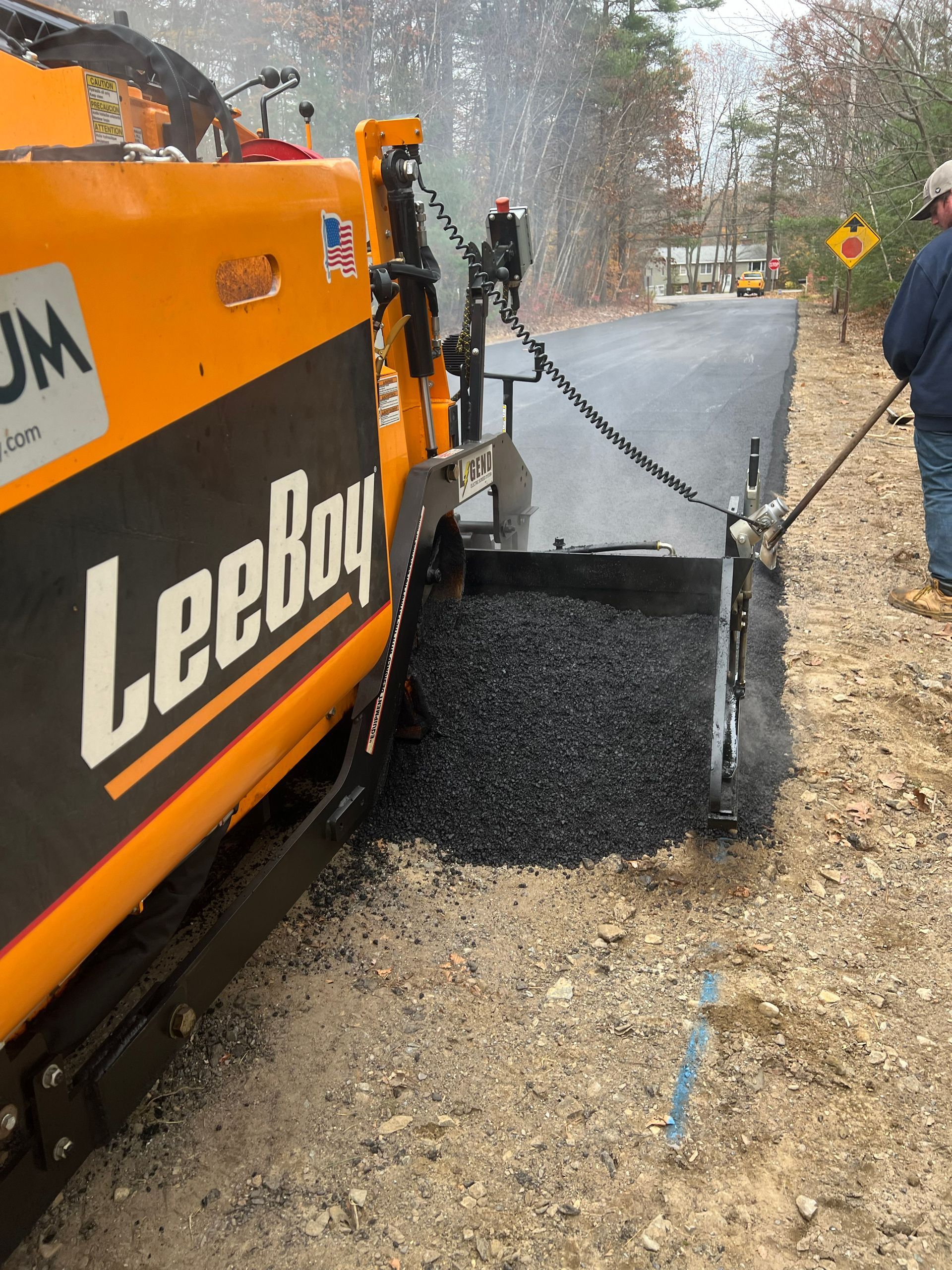 Yellow LeeBoy paving machine laying asphalt on a road. Worker stands nearby, controlling flow.