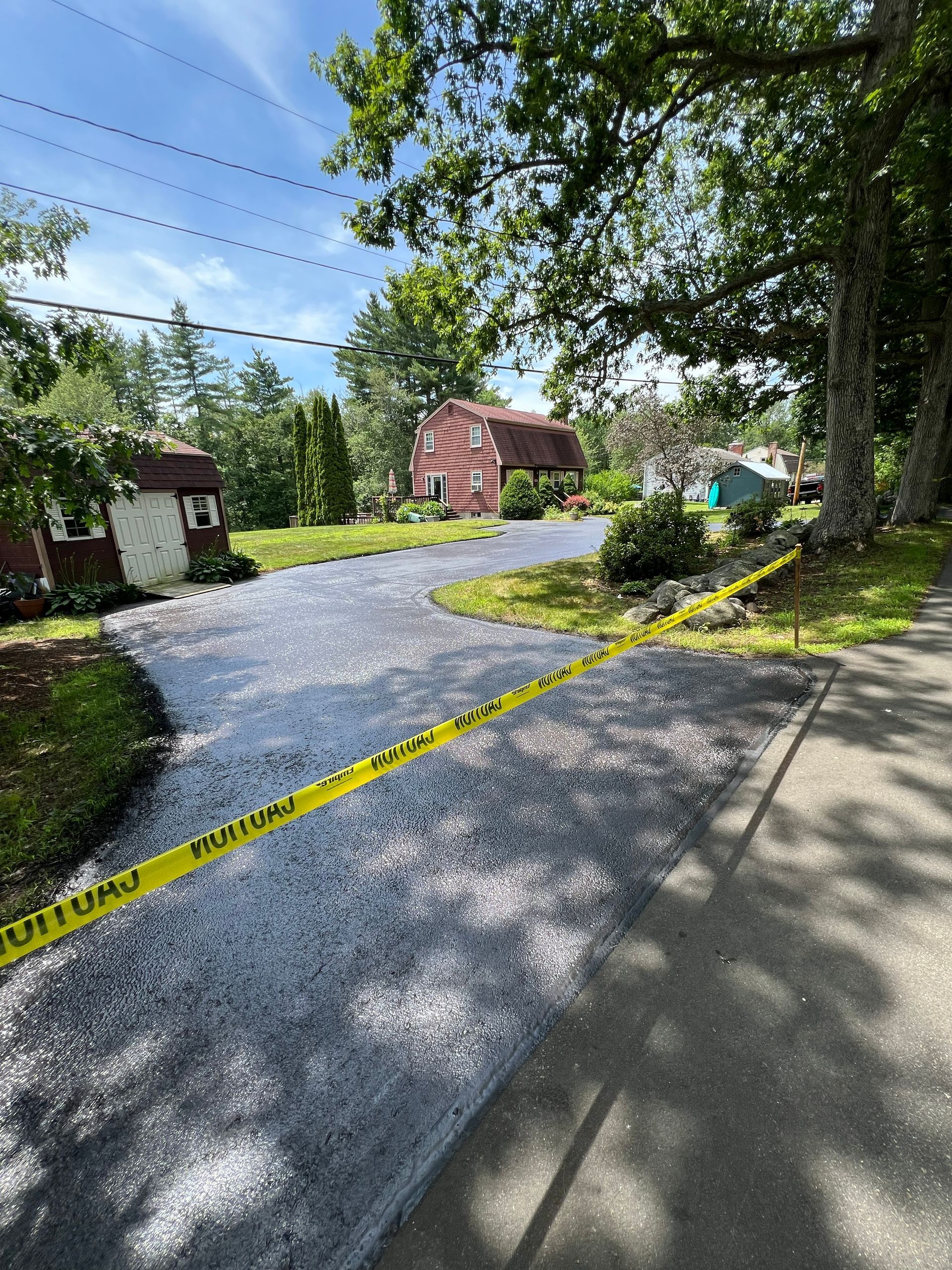 New asphalt driveway with caution tape. House and trees in background under a blue sky.
