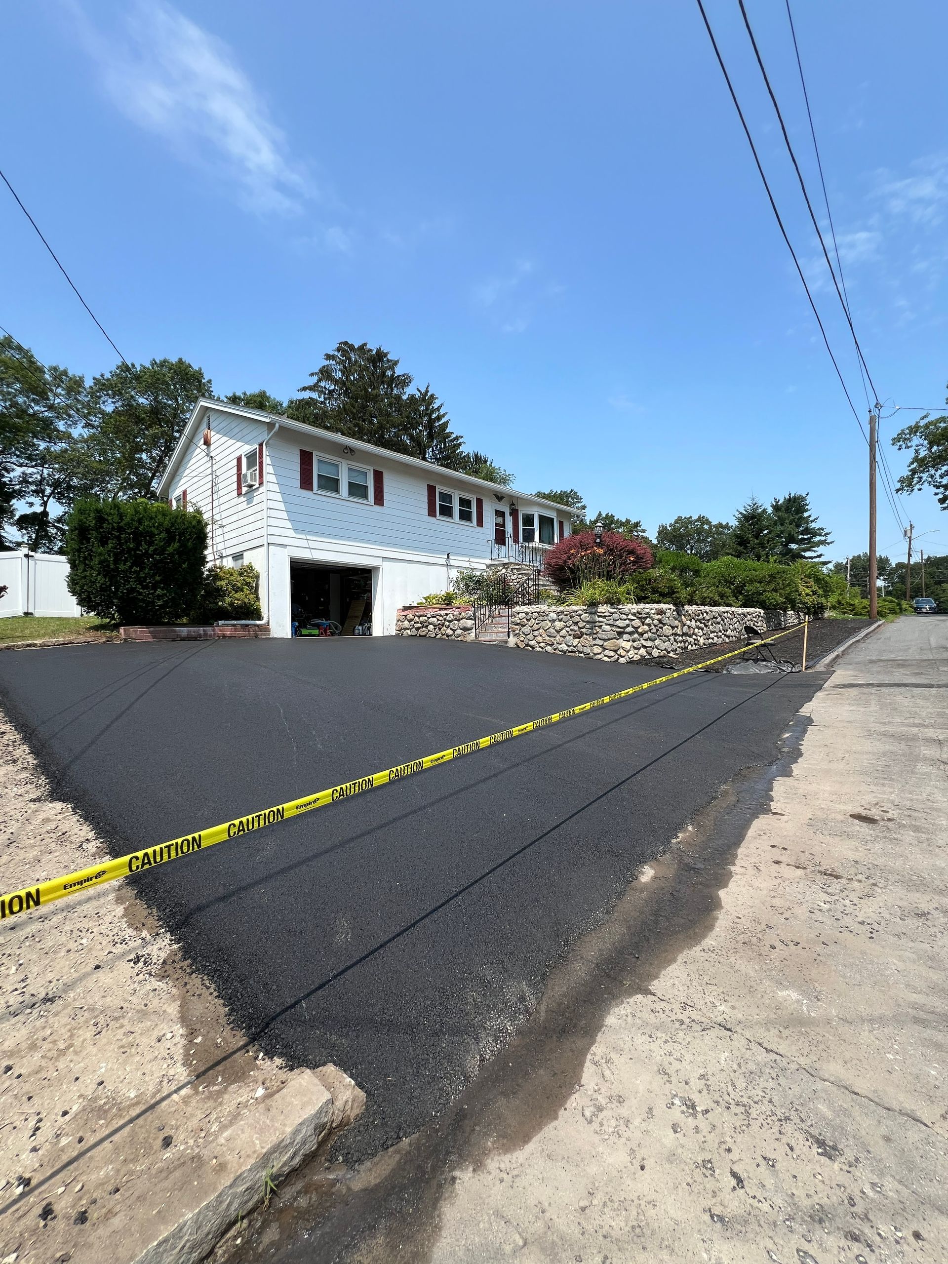 Freshly paved driveway in front of a white house, with caution tape and utility poles overhead.