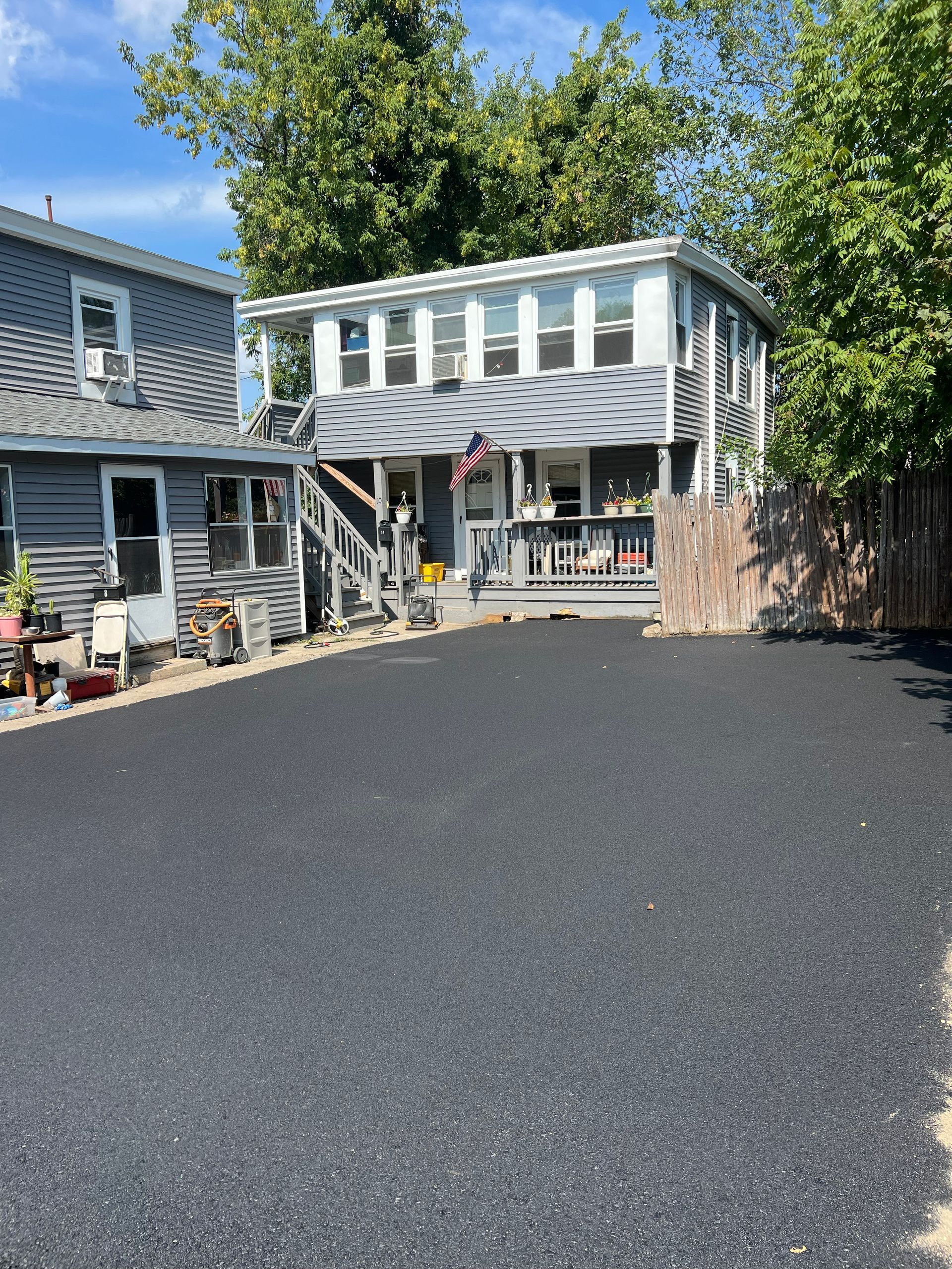 Two-story house with a newly paved driveway. Gray siding, white trim, and trees on a sunny day.