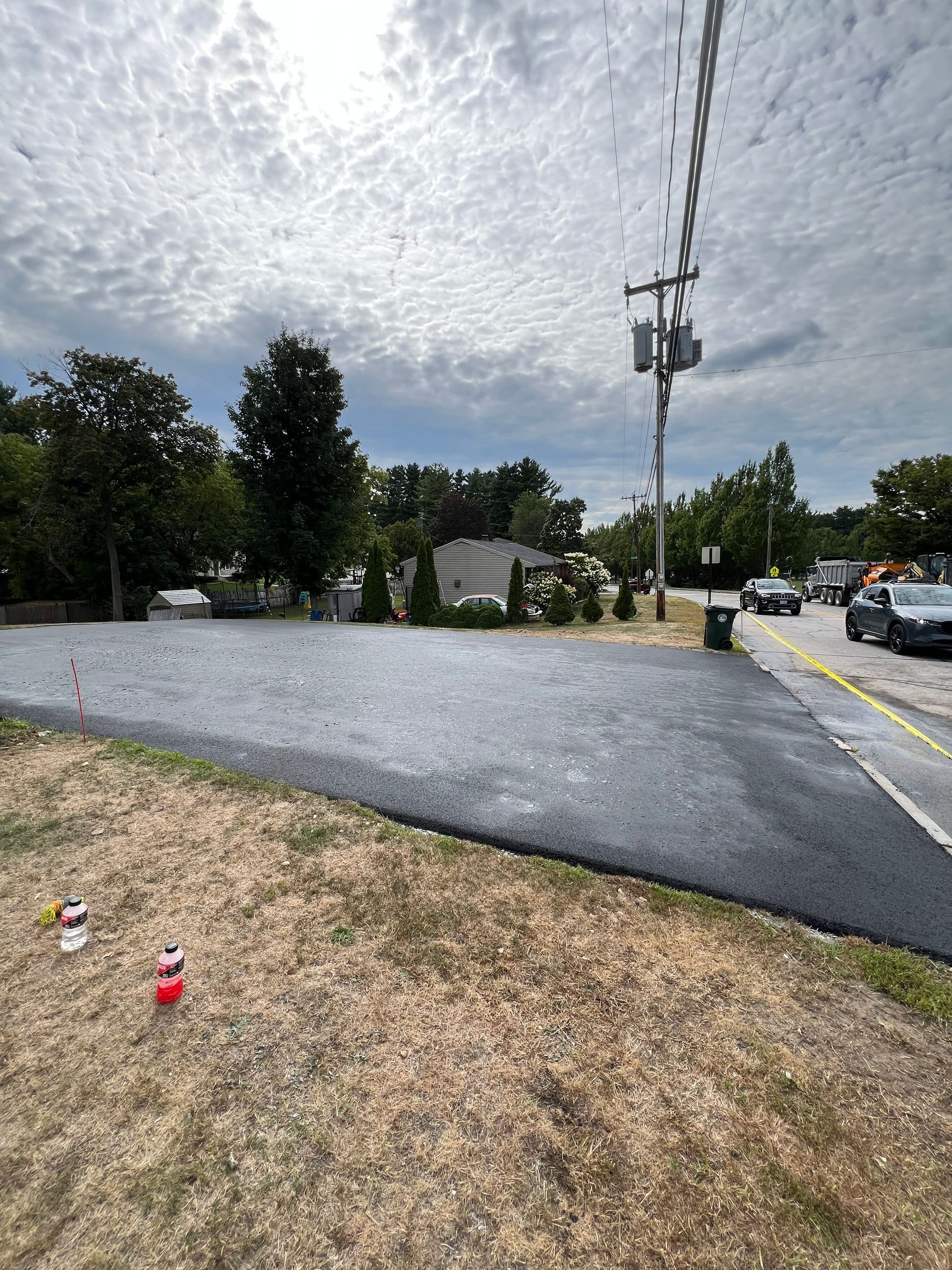 New asphalt patch next to grassy area, road with vehicles, power lines, and cloudy sky.