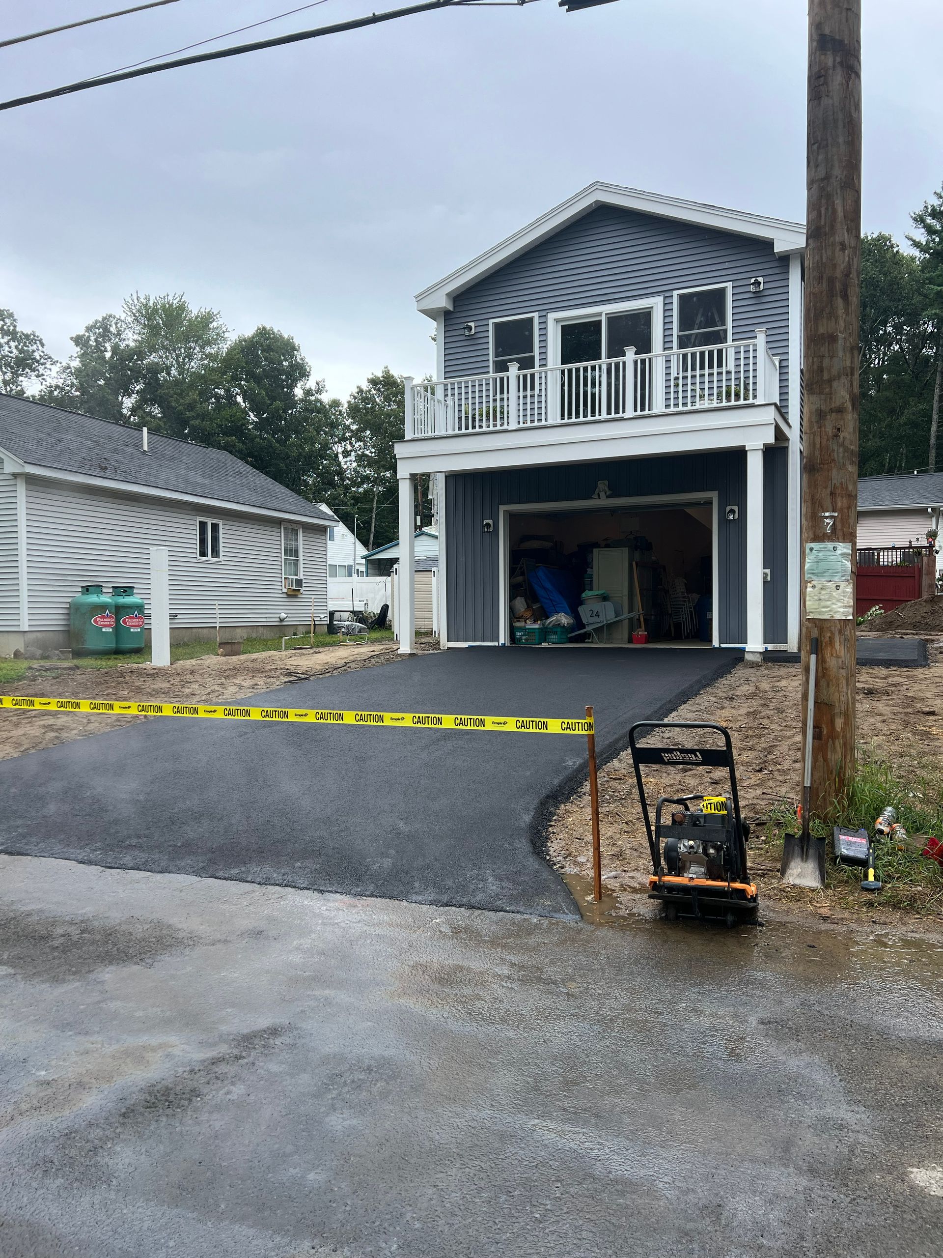 Two-story blue house with open garage. New asphalt driveway with caution tape. Compactor machine visible.