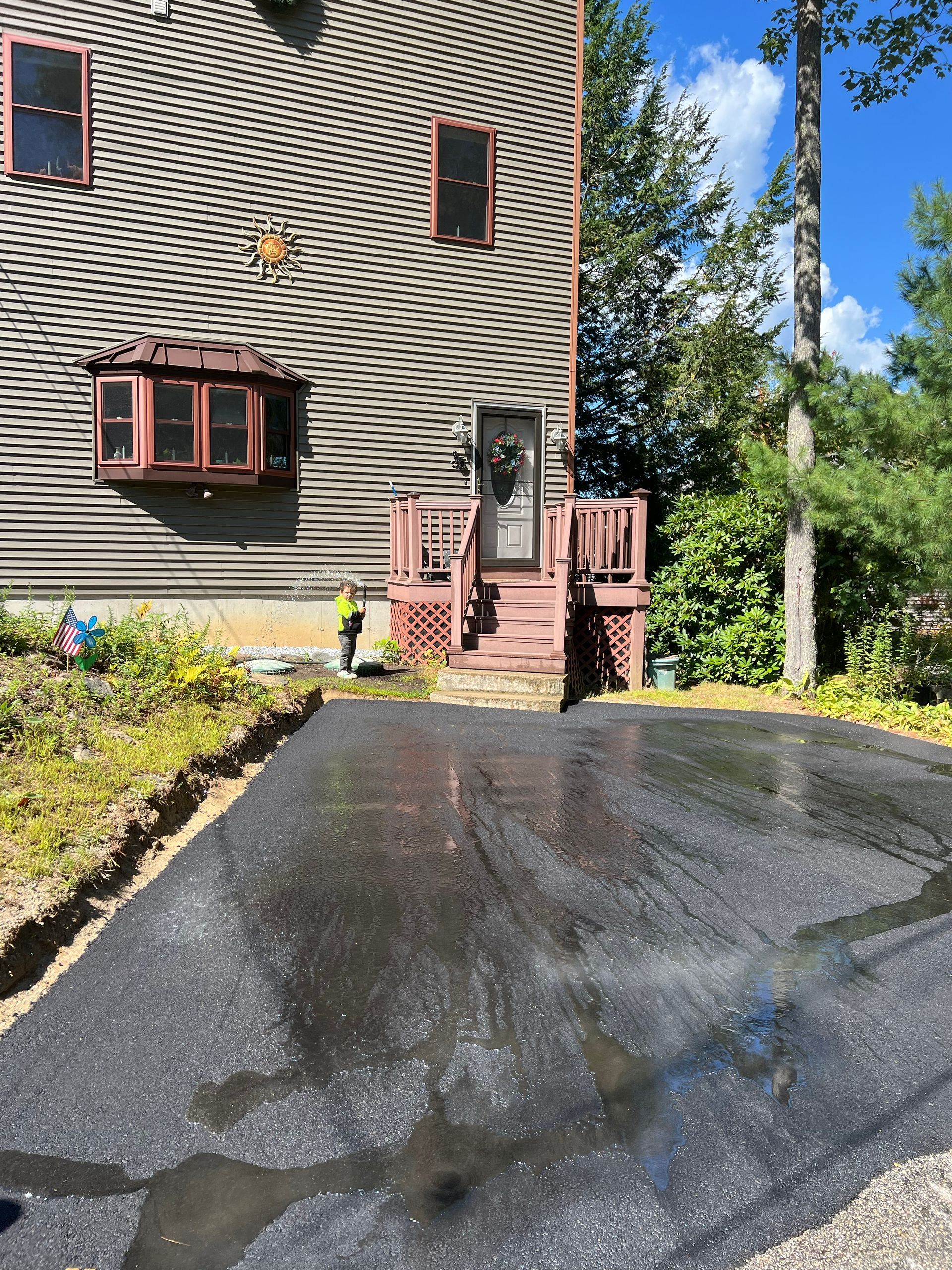 Blacktop driveway leads to a house with a brown roof and a small deck with a door.