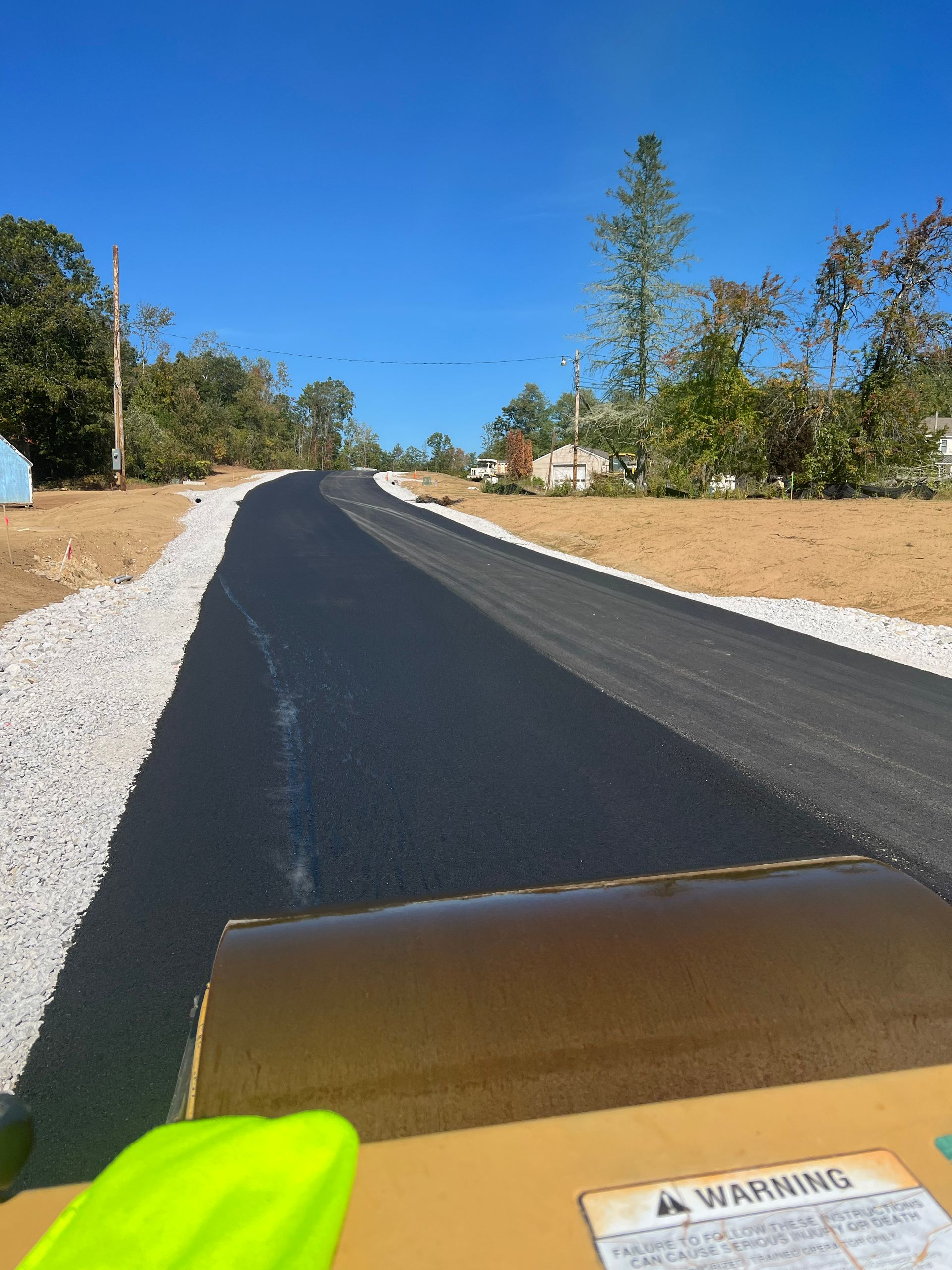 Freshly paved asphalt road, view from roller, bordered by gravel and trees under a clear blue sky.