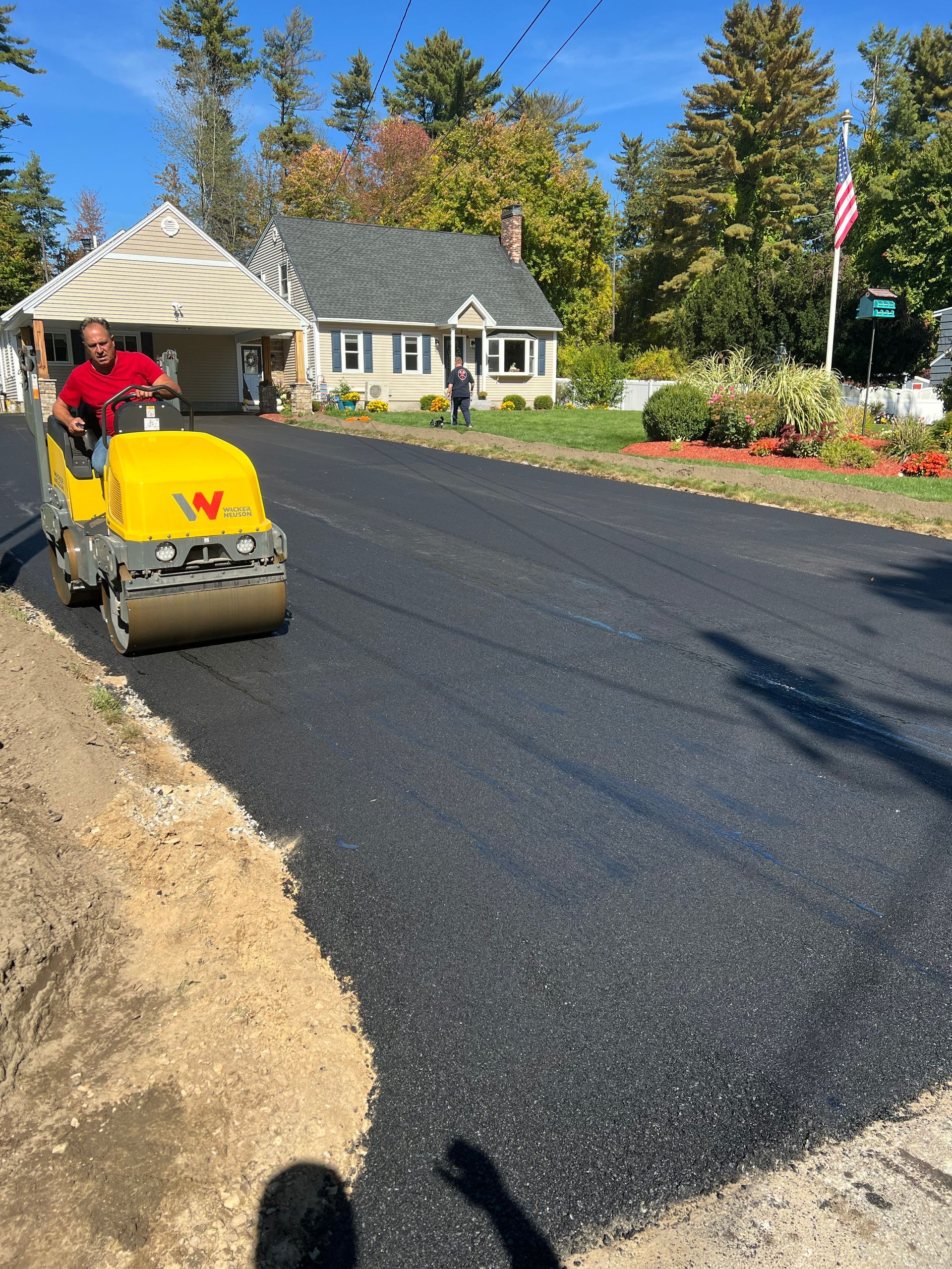 Asphalt paving a road in front of a house. A worker operates a roller compactor on fresh asphalt.