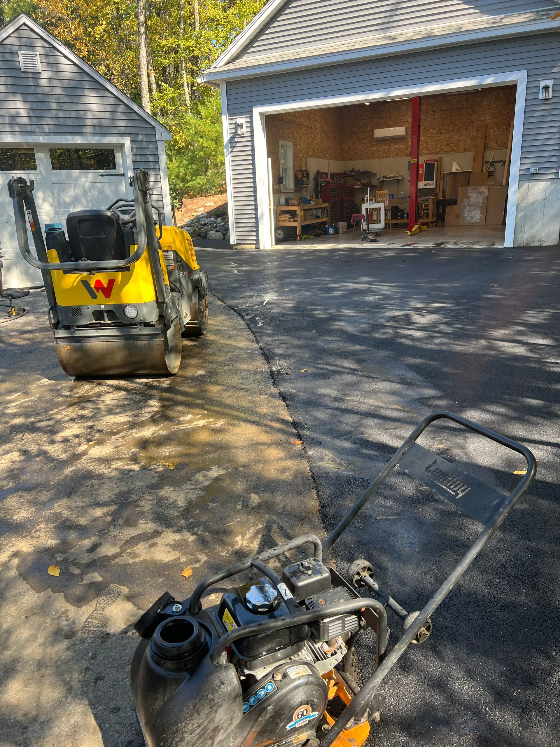 Asphalt paving in progress: Roller and plate compactor compacting black asphalt driveway near a garage.