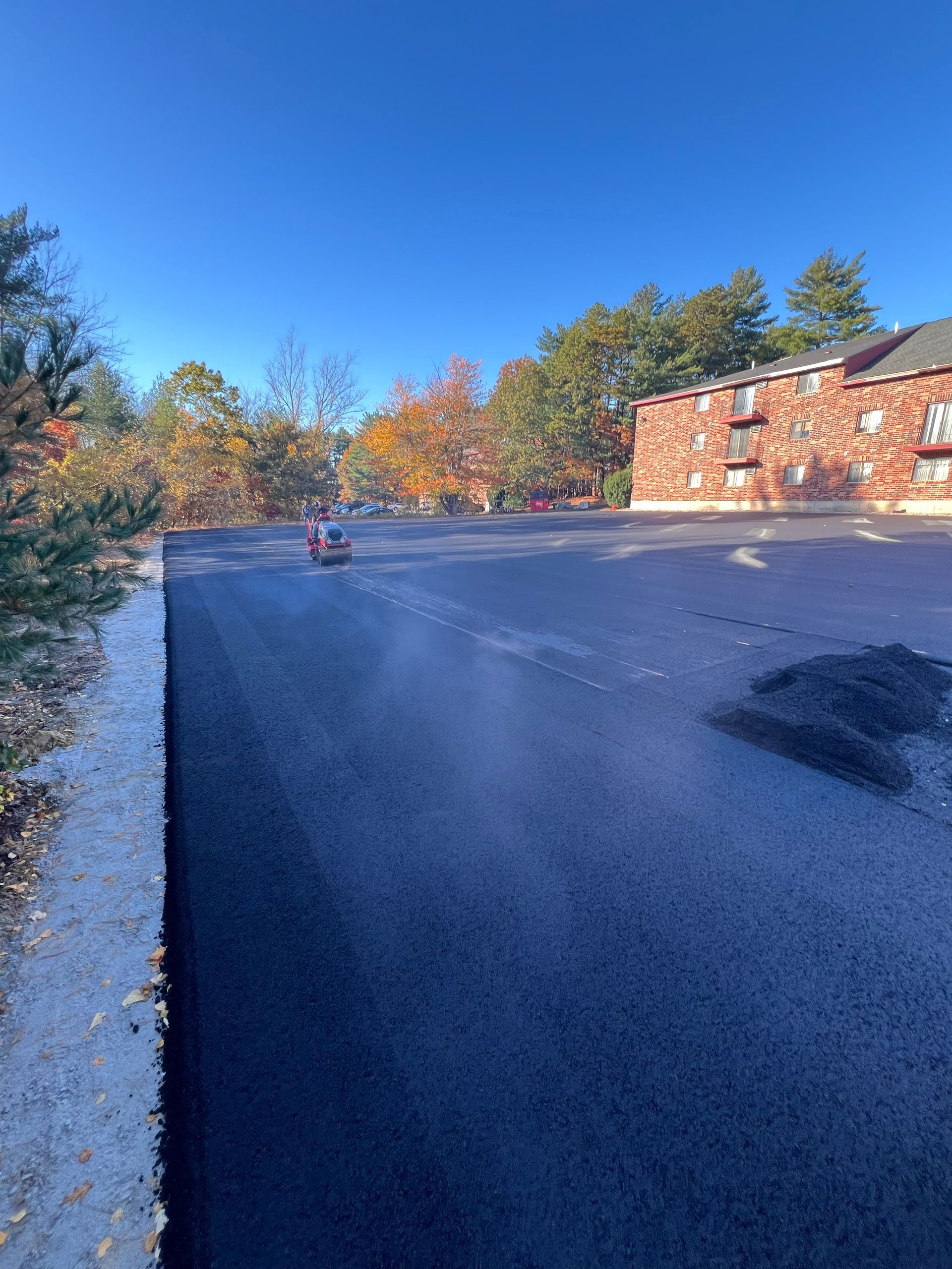 Newly paved asphalt parking lot with a brick building in the background. Bright blue sky with trees.