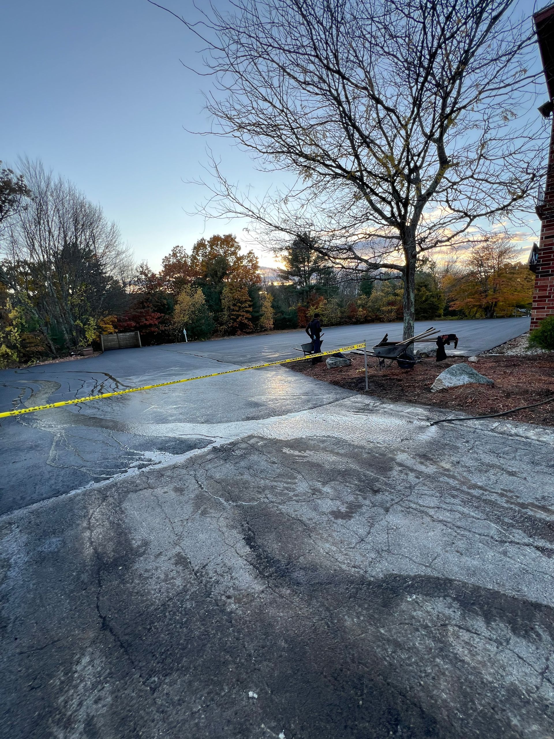 Asphalt driveway with yellow caution tape, tree, and distant view of trees under a blue sky.