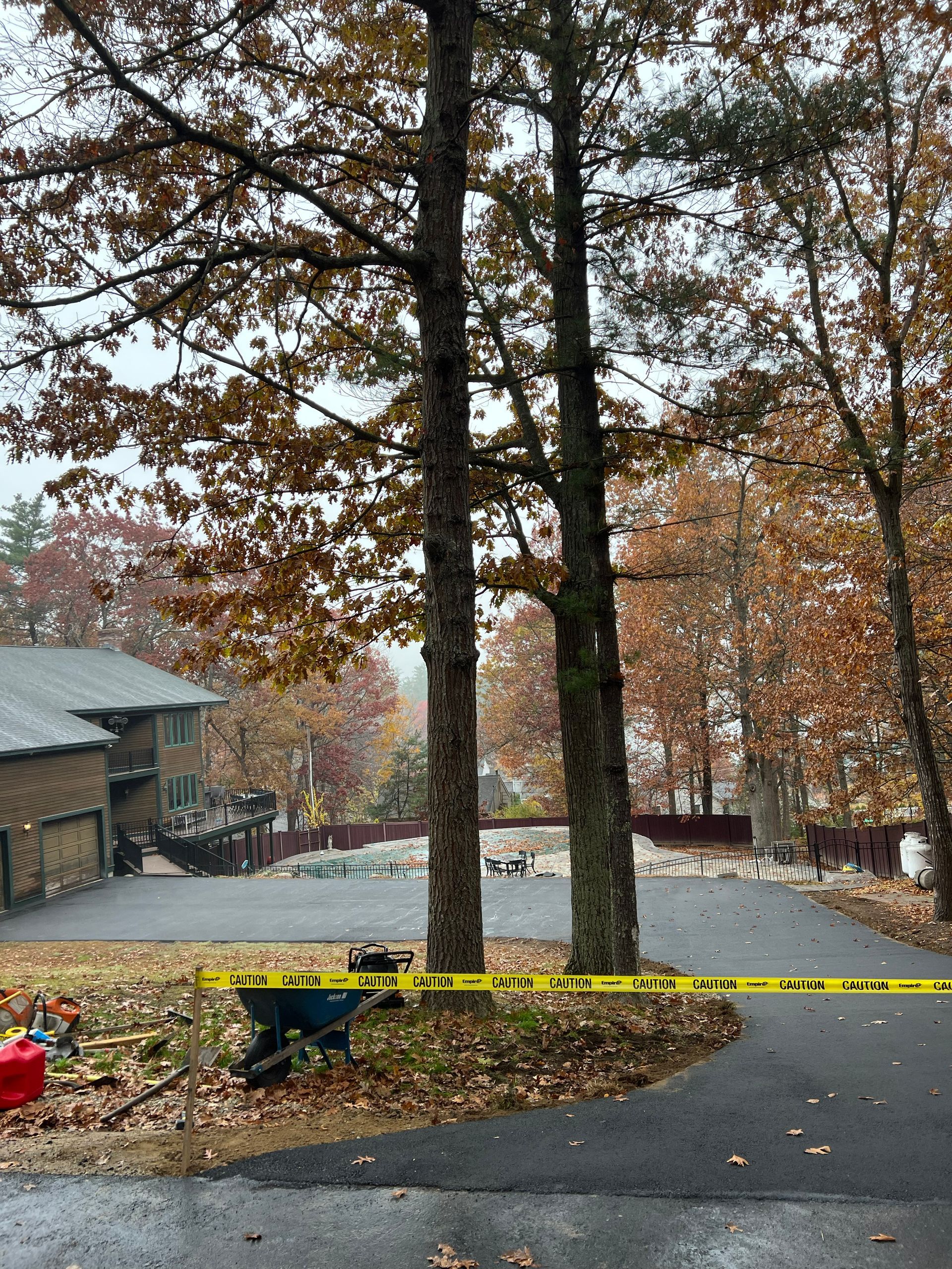 Two tall trees flank a driveway cordoned off with yellow tape; a house and fall foliage are in the background.