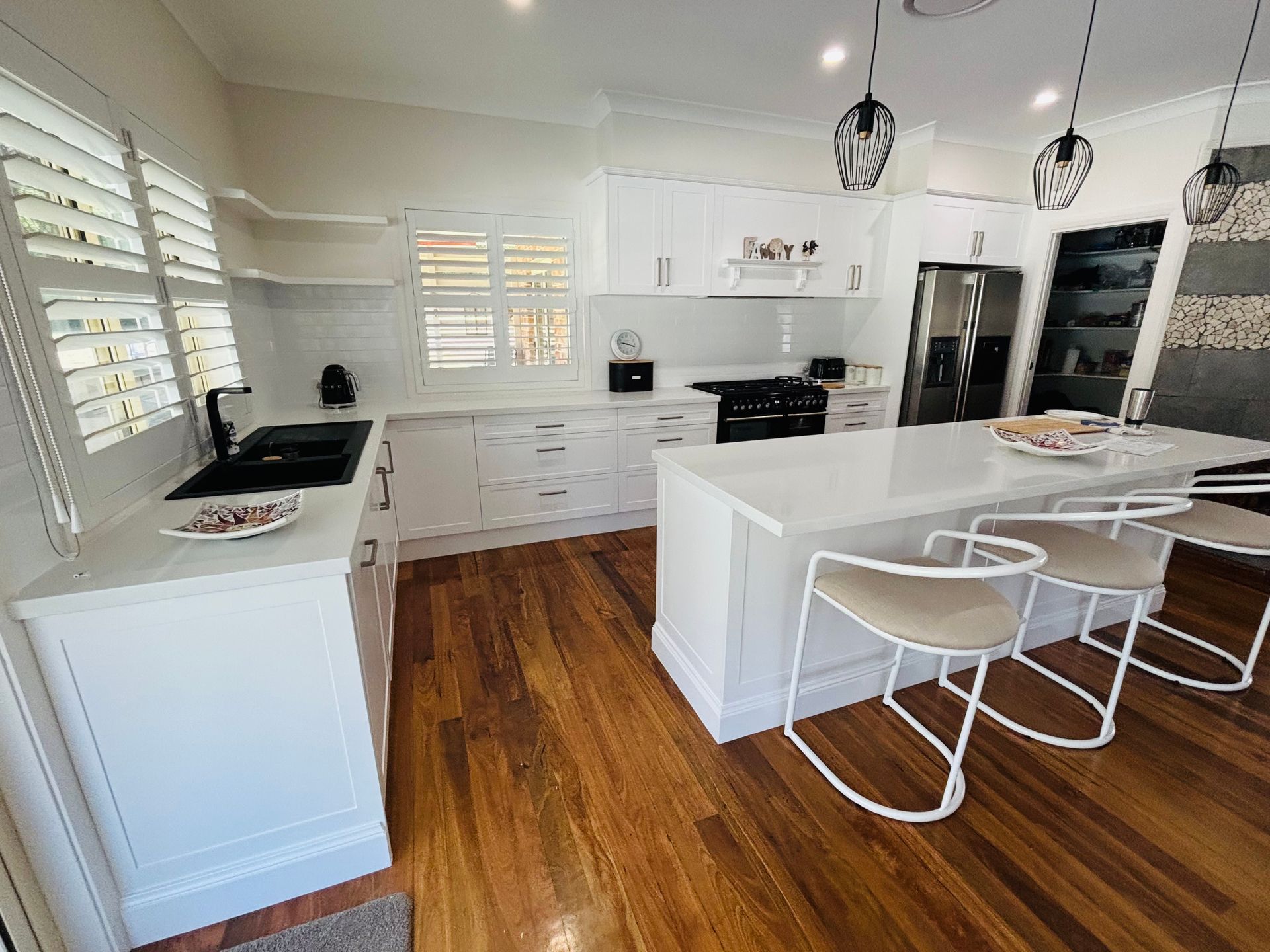White kitchen with island, cabinets, dark appliances, and wood floor. Shutters and pendant lights. — Piscopo Kitchens Plus In Forster, NSW