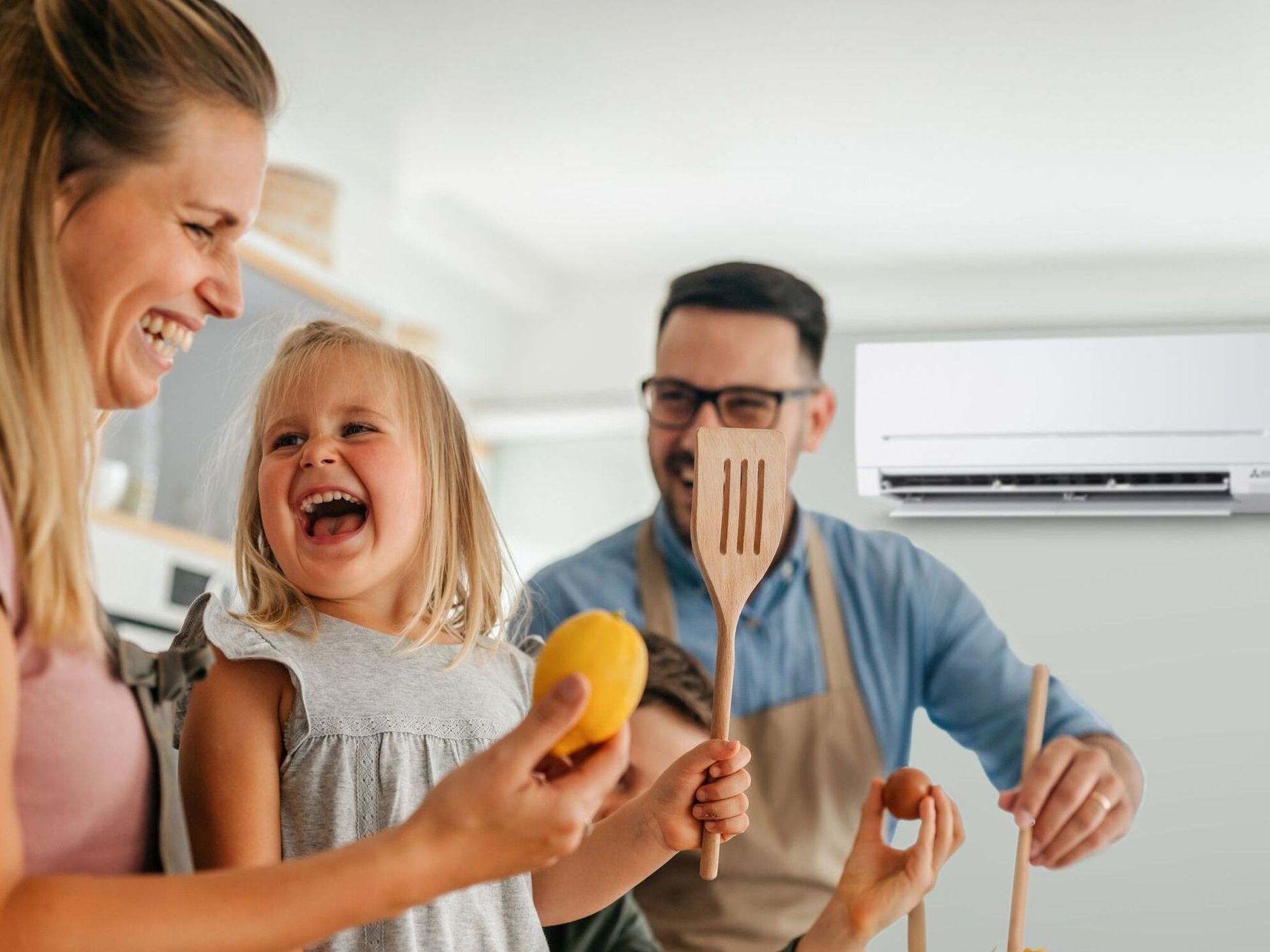A family laughs while cooking in a bright kitchen. An air conditioner is mounted on the wall.
