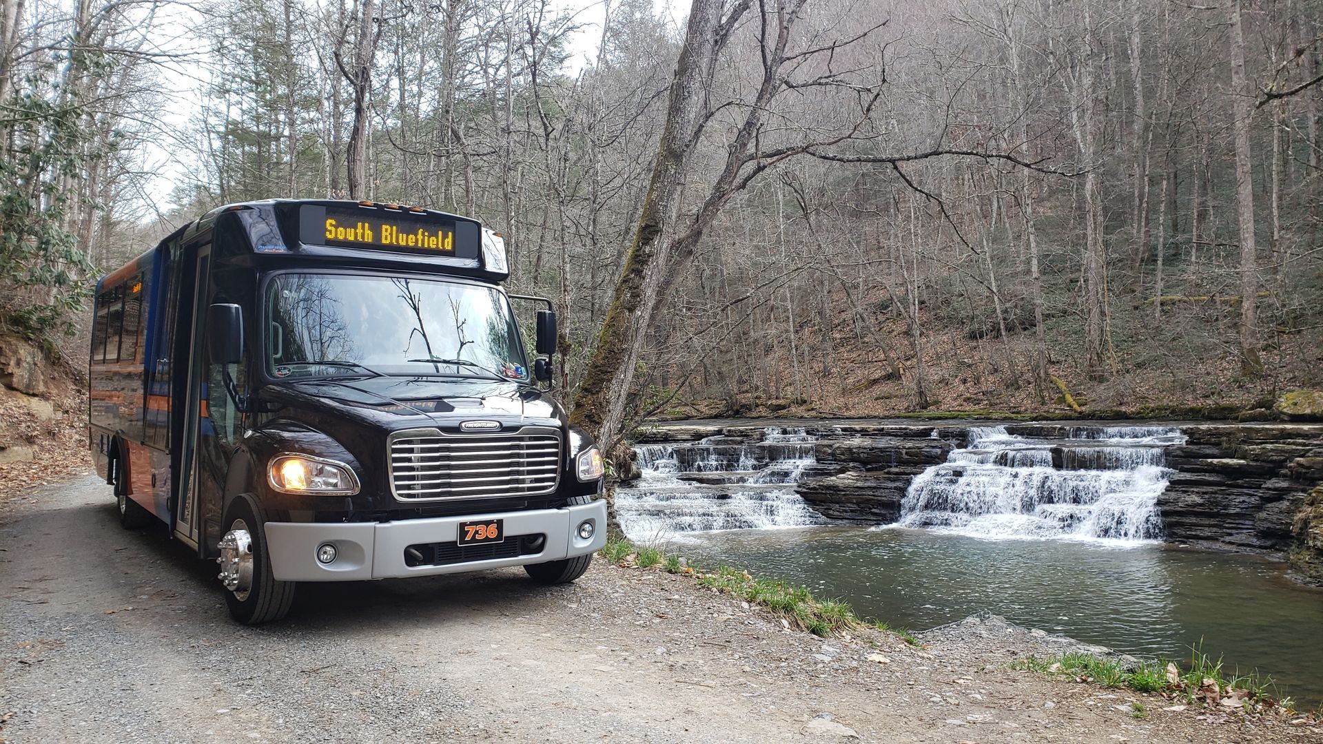 Black shuttle bus parked on gravel road beside small waterfall in wooded area.