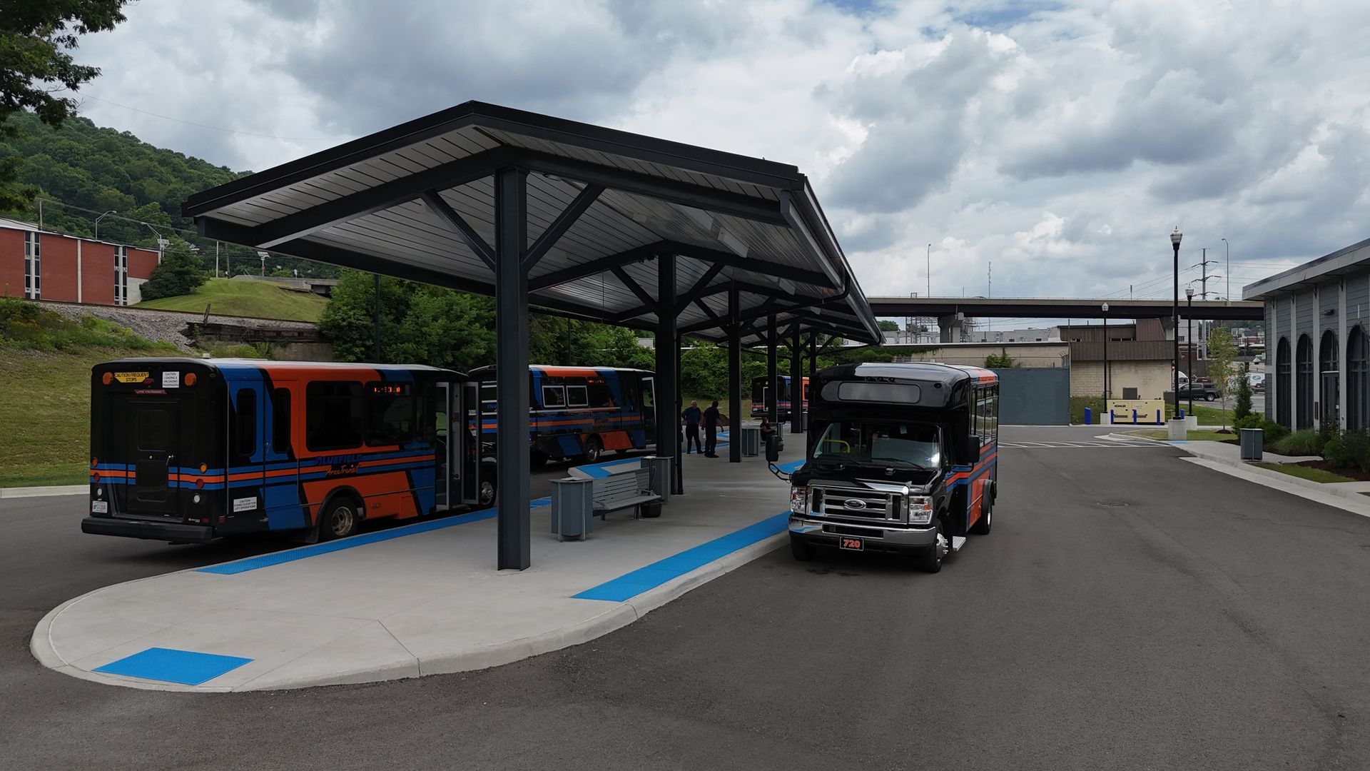 Three buses at a bus stop under a shaded roof. Cloudy sky, building in background.