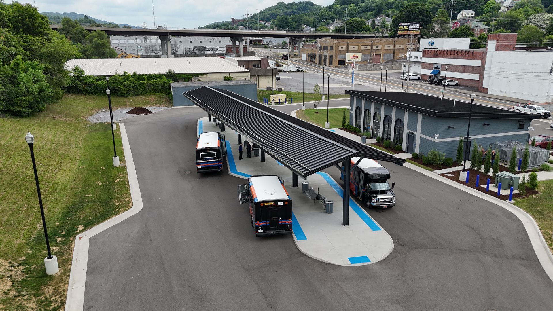 Outdoor transit station with buses under a dark canopy; buildings and bridge in the background.