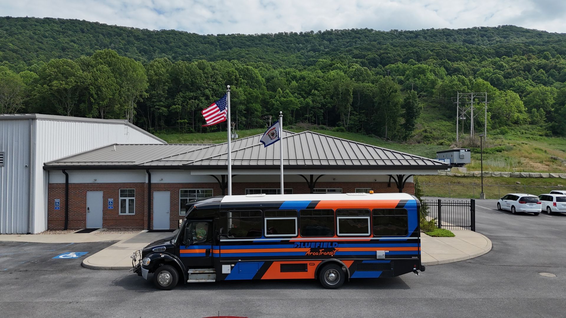 A black and blue shuttle bus in front of a building with American and state flags. A mountain is in the background.