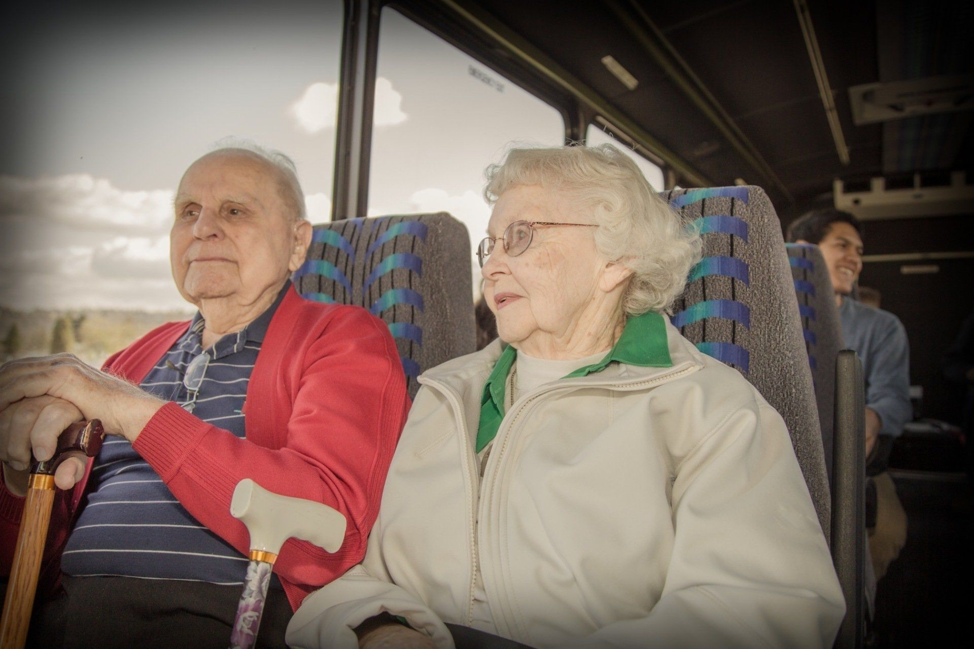 Elderly couple sitting on a bus; man with cane, woman in glasses, looking out the window.